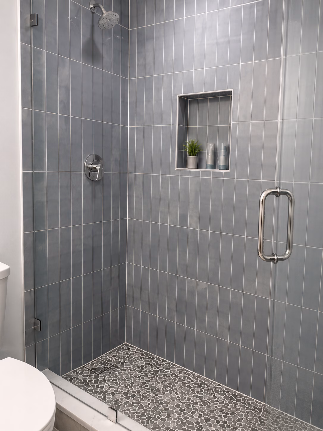 Slate-blue vertical tile shower with a niche, glass door and pebble floor, remodeled by BFM Builders